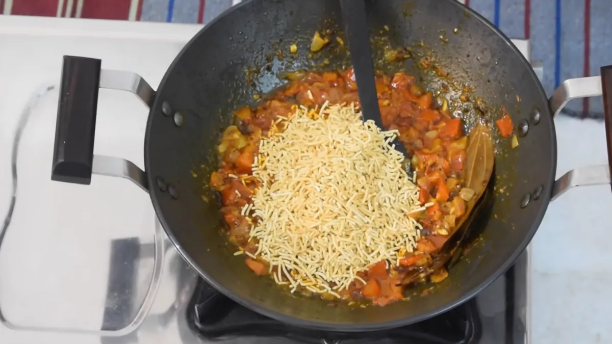 Kadhai me pak rahi tamatar ki gravy par sev daali ja rahi hai